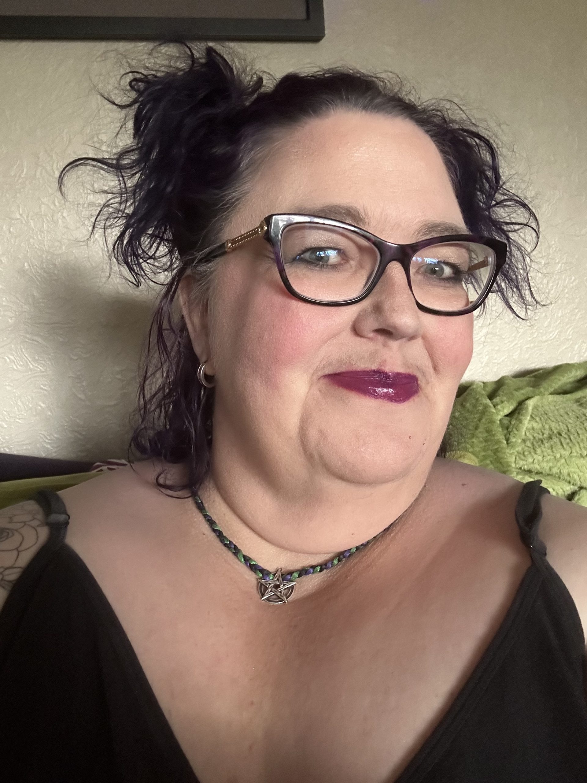 a fat white woman with her dark purple (almost black) hair pinned up to either side, wearing black-and-purple glasses and berry lipstick. I am wearing a pentacle necklace and have a spaghetti-strap top on.