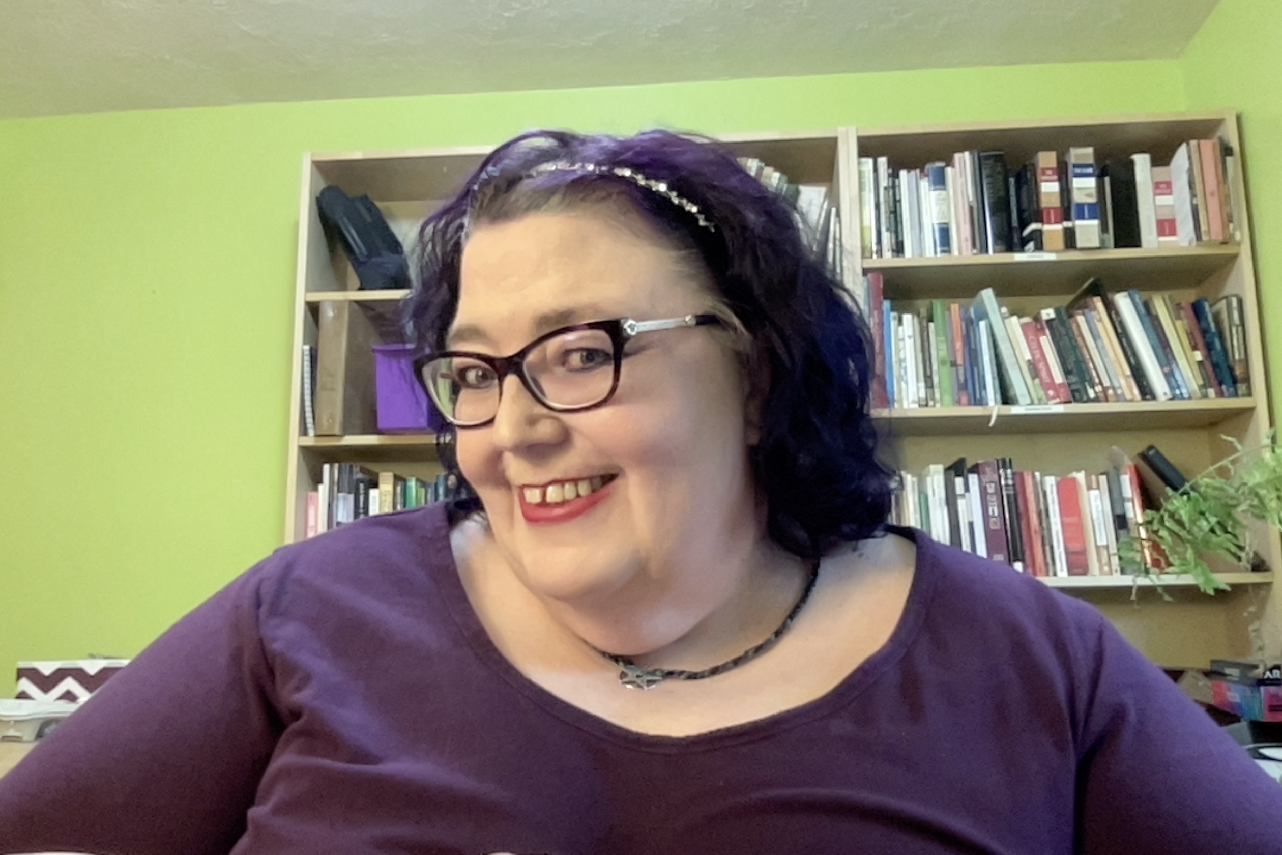 A fat white femme with a sparkly silver headbad over her shoulder-length dark violet hair. She is wearing a somewhat lighter violet dress with a scoop neckline. There are bookcases behind her.